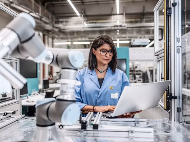 A person standing in a factory working on a laptop. She is surrounded by robot arms and machinery.