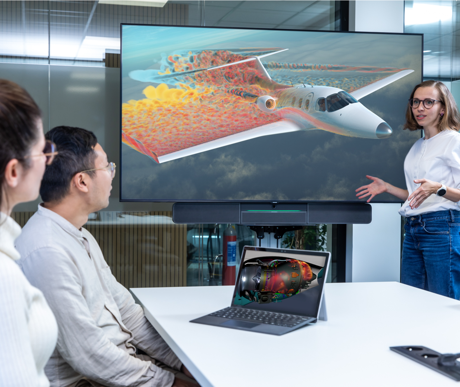 Three people gathered in a meeting room with a large screen displaying a simulation of a plane flying. One is presenting while the others are sitting down and listening.